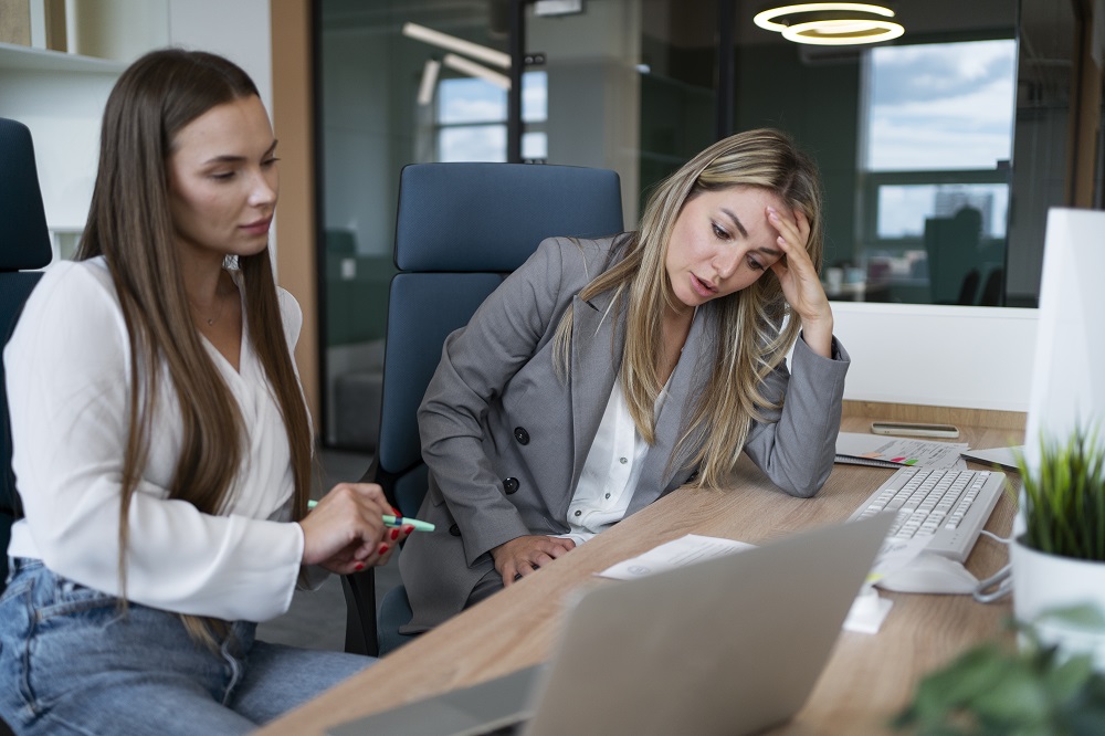 Duas mulheres estão sérias em um escritório, a do lado direito está com a mão na cabeça e expressão preocupada. 