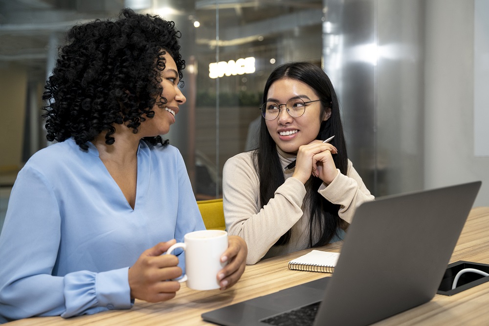 Duas mulheres estão em um escritório conversando. Estão em frente a uma mesa com notebook e uma agenda.