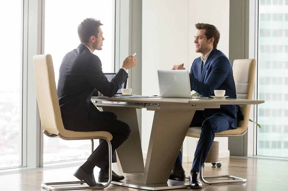 Dois homens estão sentados de frente um para o outro conversando. A imagem representa a entrevista para descrição de cargo. 