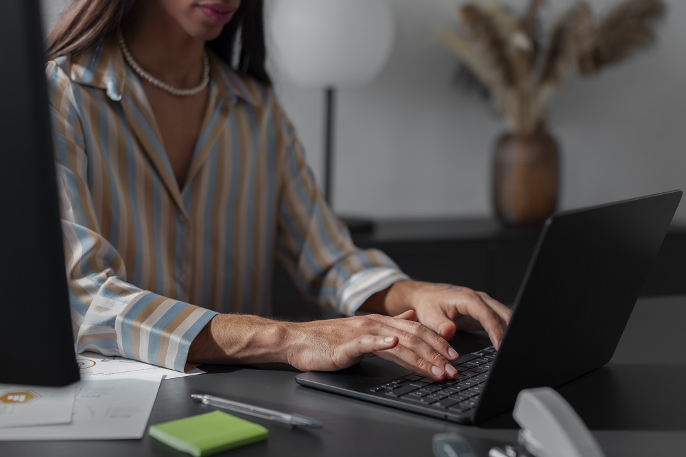 A mulher representa alguém elaborando a descrição de cargos em um notebook que está na mesa ao lado de um bloco de post-it verde e uma caneta. 