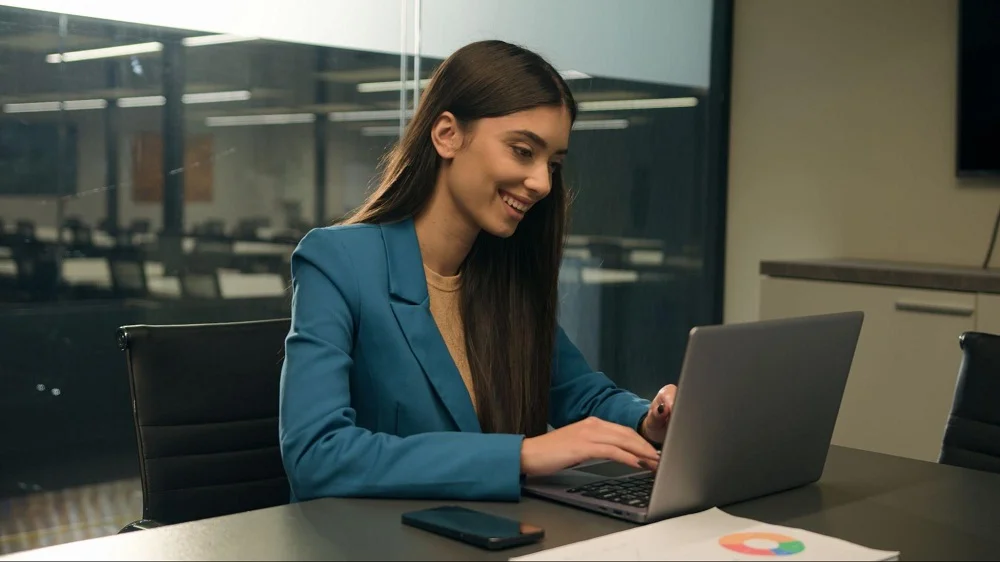 Mulher sorridente está sentada em frente a um notebook, ao fundo existe outra mesa com cadeiras. A imagem representa uma pessoa fazendo a descrição de cargos. 