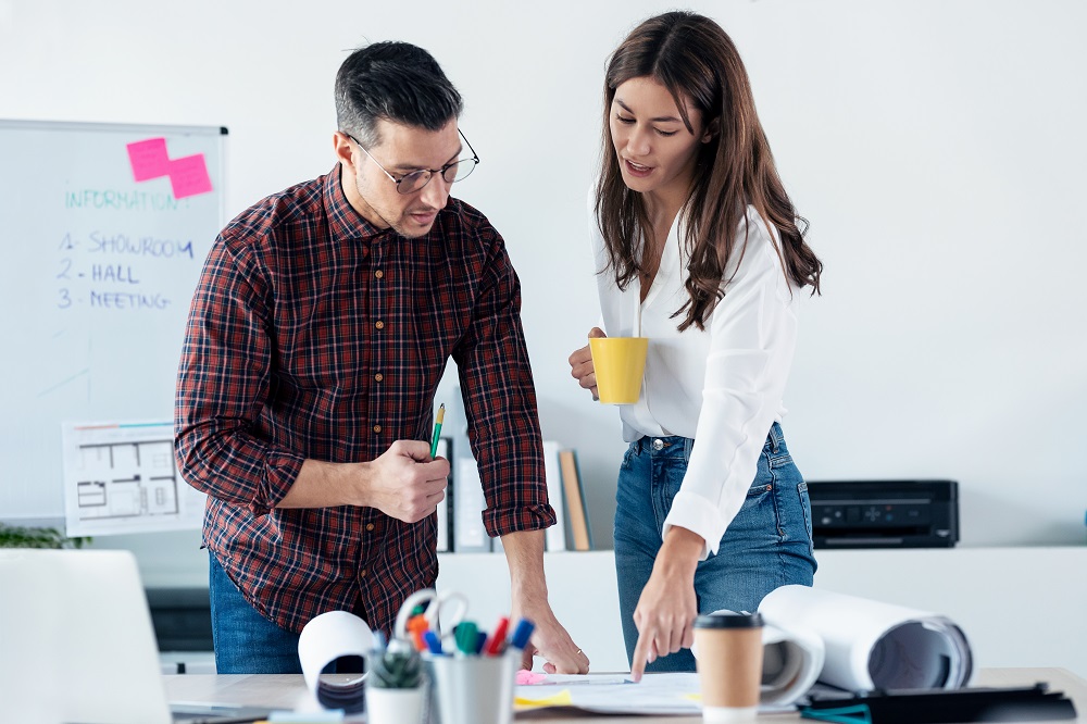 Um homem segurando um lápis e uma mulher segurando uma caneca amarela estão em frente a uma mesa apontando para alguns documentos. Ao fundo existe uma lousa branca com post-it rosa e algumas coisas escritas em preto.