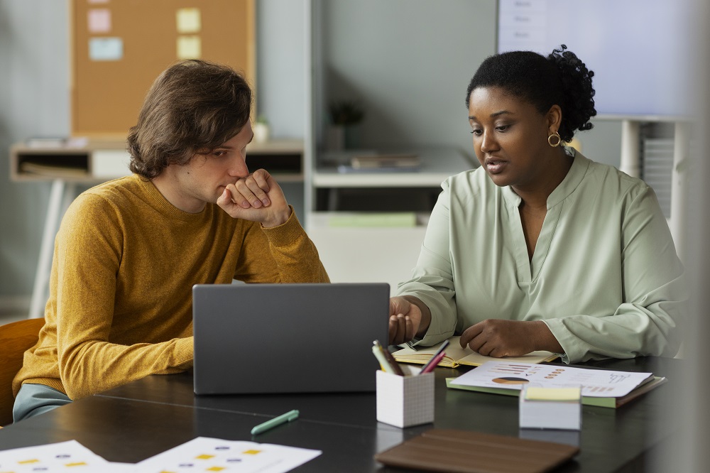 Um homem e uma mulher estão em um escritório, na mesa há um computador, alguns papéis e canetas. A imagem representa uma reunião de PDI.