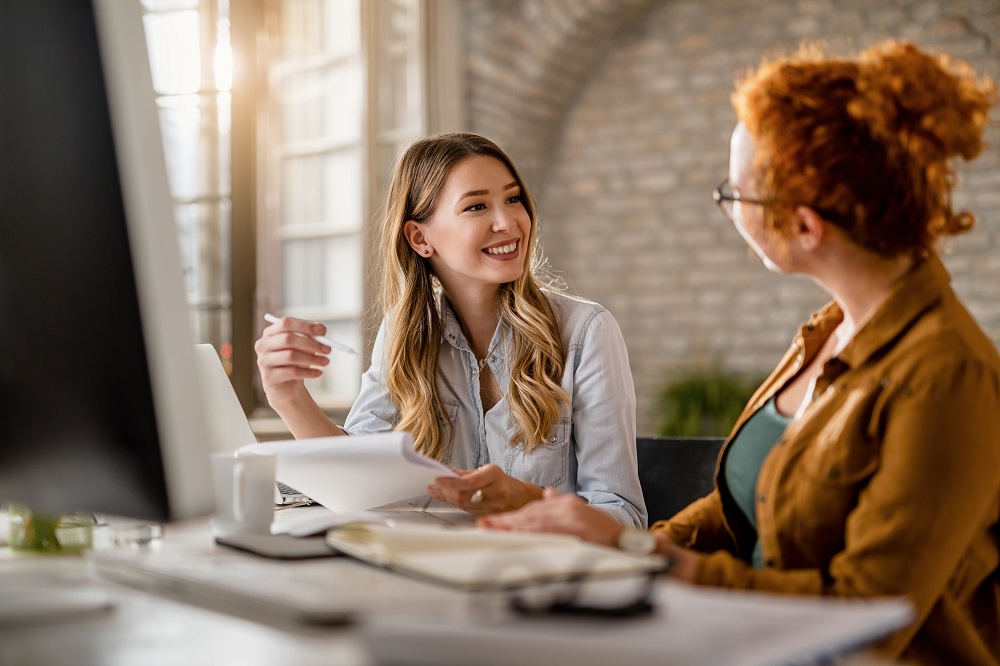 Duas mulheres sorridentes estão em uma sala em frente a um computador. A imagem representa uma reunião de alinhamento do PDI