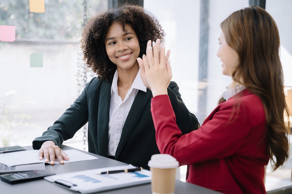 Duas mulheres sorriem sentadas em frente a uma mesa, estão batendo a mão uma na outra como se estivessem comemorando