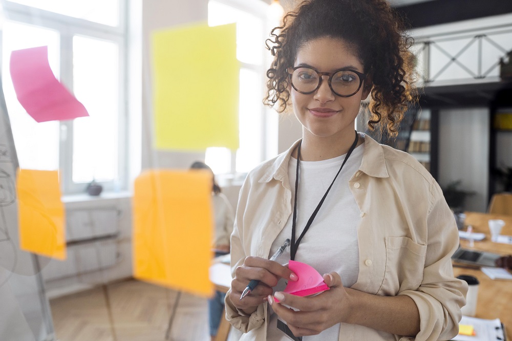 Na imagem há uma mulher sorrindo enquanto olha para frente de uma porta de vidro com quatro post-it colados