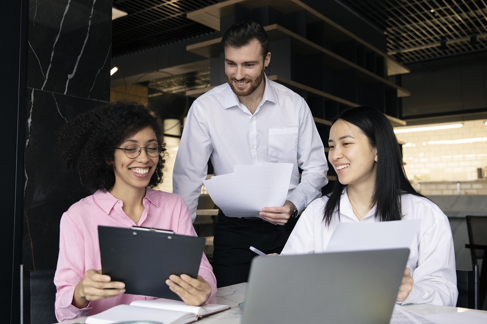 Na imagem tem três pessoas sorridentes em um escritório, estão olhando para uma prancheta. A imagem representa o aprendizado em equipe.