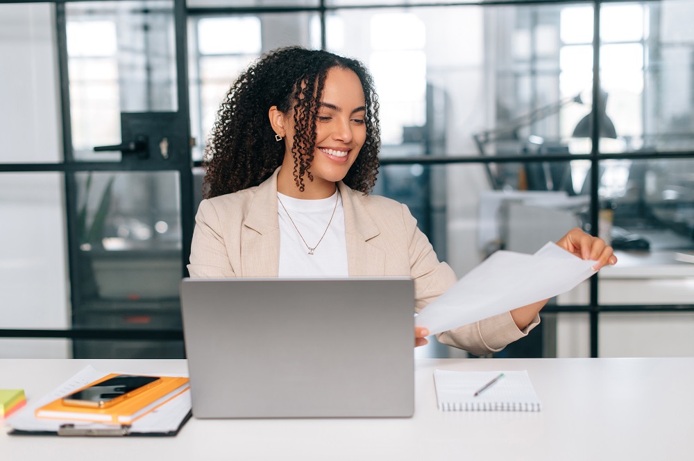 Mulher de cabelos cacheados está sorrindo enquanto olha alguns papéis em sua mão. A sua frente, há um notebook aberto e uma agenda laranja com um celular em cima.