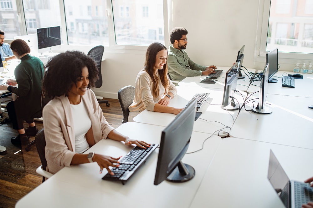 Equipe trabalhando em um escritório, utilizando plataforma LMS em computadores, com ambiente iluminado por luz natural.