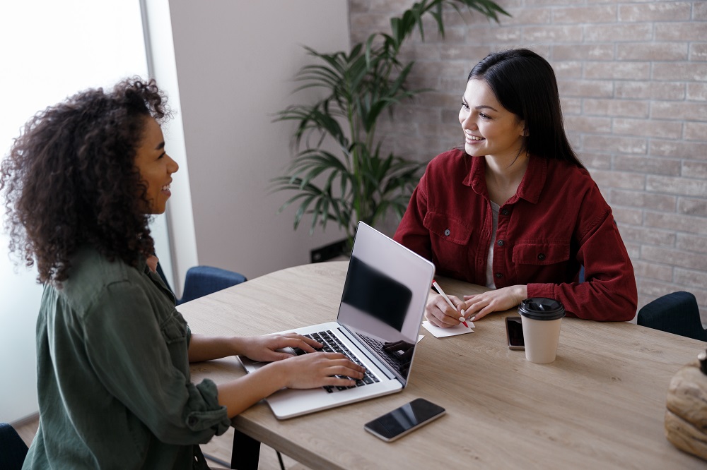 Duas mulheres sorridentes estão em uma sala, uma delas está mexendo no notebook, enquanto a outra faz anotações em um bloco de notas.