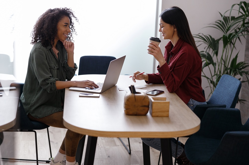 Duas mulheres estão em uma sala, uma está em frente ao notebook enquanto a outra segura um copo de café.