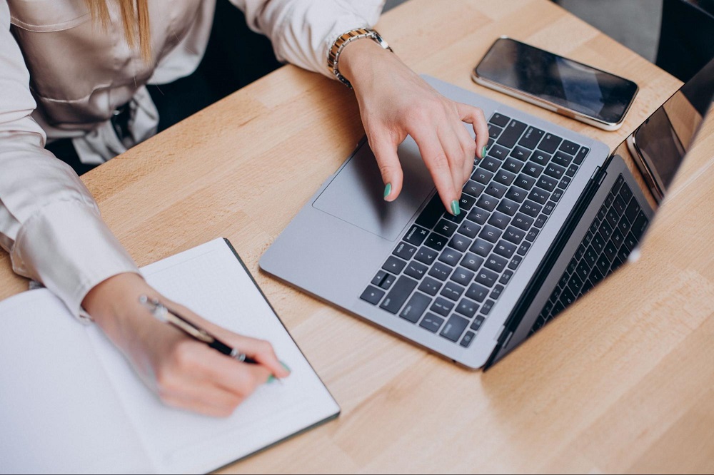  A imagem mostra as mãos de uma mulher, ela está mexendo em um notebook e anotando em uma agenda. A imagem representa a implementação do LMS no processo de onboarding. 