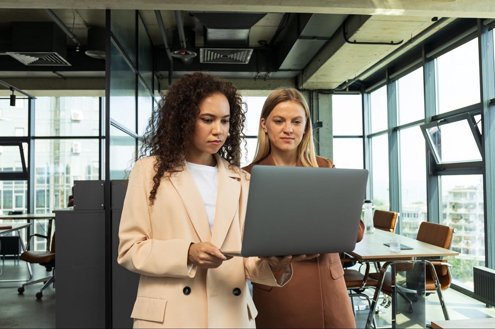 Duas mulheres estão segurando um notebook cinza. Ao fundo, existem algumas cadeiras e mesa de reunião. 