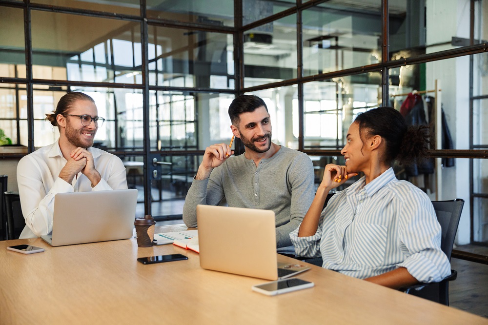 Três pessoas sorridentes estão sentadas em uma sala de reunião, existem dois notebooks na mesa ao lado de três celulares. A imagem representa uma reunião de alinhamento sobre o workflow.