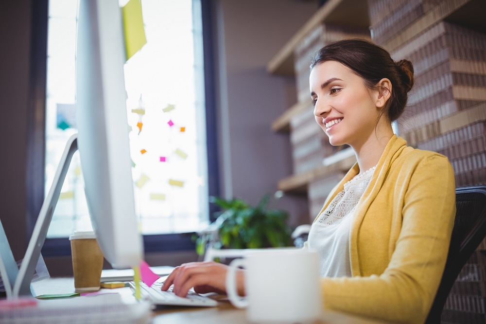 Mulher sorridente está em frente a um computador, ao lado há uma xícara branca e do outro um copo de plástico.