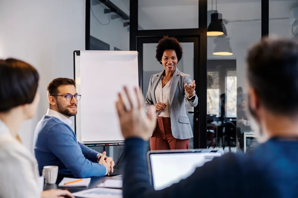 Quatro pessoas estão em uma sala de reunião, o foco está em uma mulher ao lado de um quadro em branco e um homem levantando a mão para fazer uma pergunta. A imagem representa uma conversa para o treinamento corporativo.