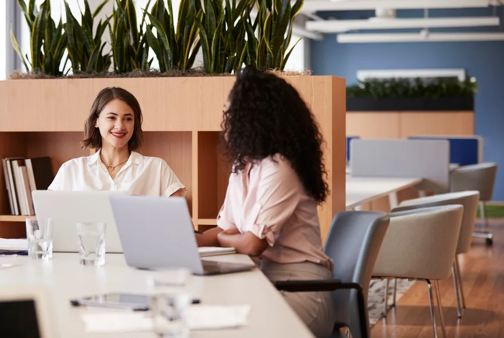 Duas mulheres estão sentadas em frente a um computador, uma delas está sorrindo enquanto olha para a outra. A imagem representa um dos tipos de feedback.