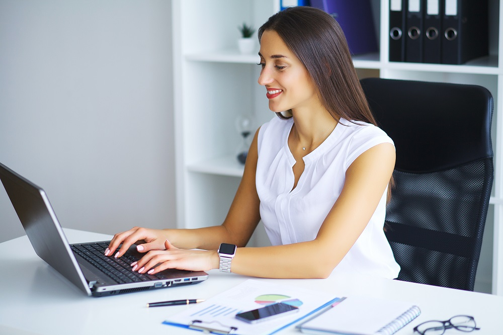 Uma mulher está sentada em frente a um notebook, na mesa há alguns papéis e caneta. 