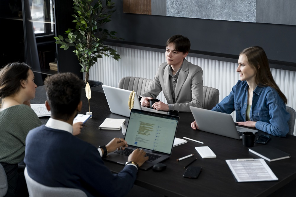 Uma equipe de 4 pessoas está reunida em uma sala de reuniões, cada um está em frente de um notebook. A imagem representa uma reunião estratégica para falar sobre Balanced Scorecard. 