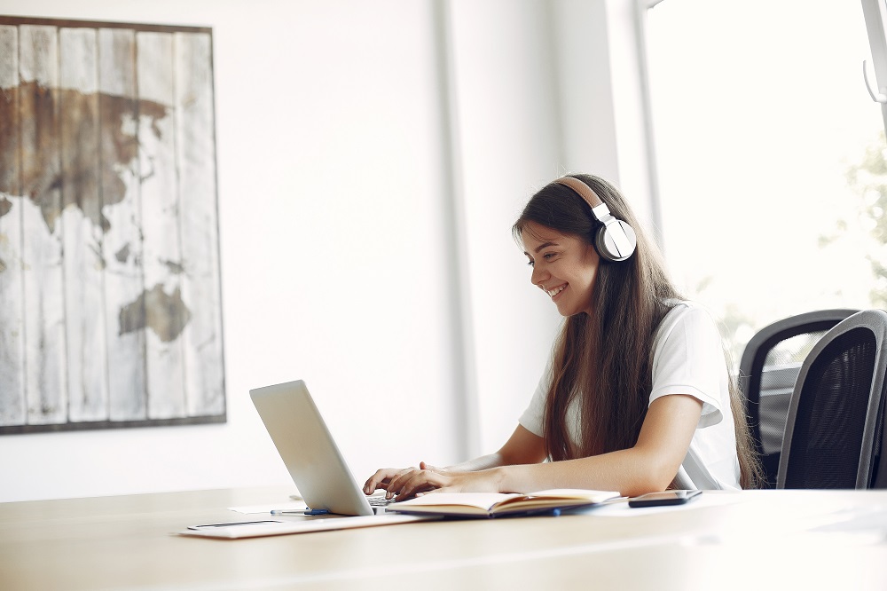 Uma mulher está de fone sorrindo para o notebook, na mesa há um caderno aberto e um celular. 