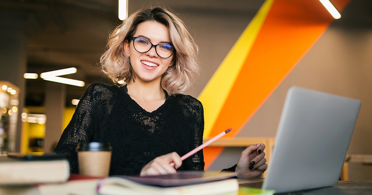 Uma mulher jovem sorridente, usando óculos, sentada à mesa de um escritório moderno com um notebook aberto e um copo de café, representando a análise dos quadrantes da matriz 9 box na gestão de talentos.