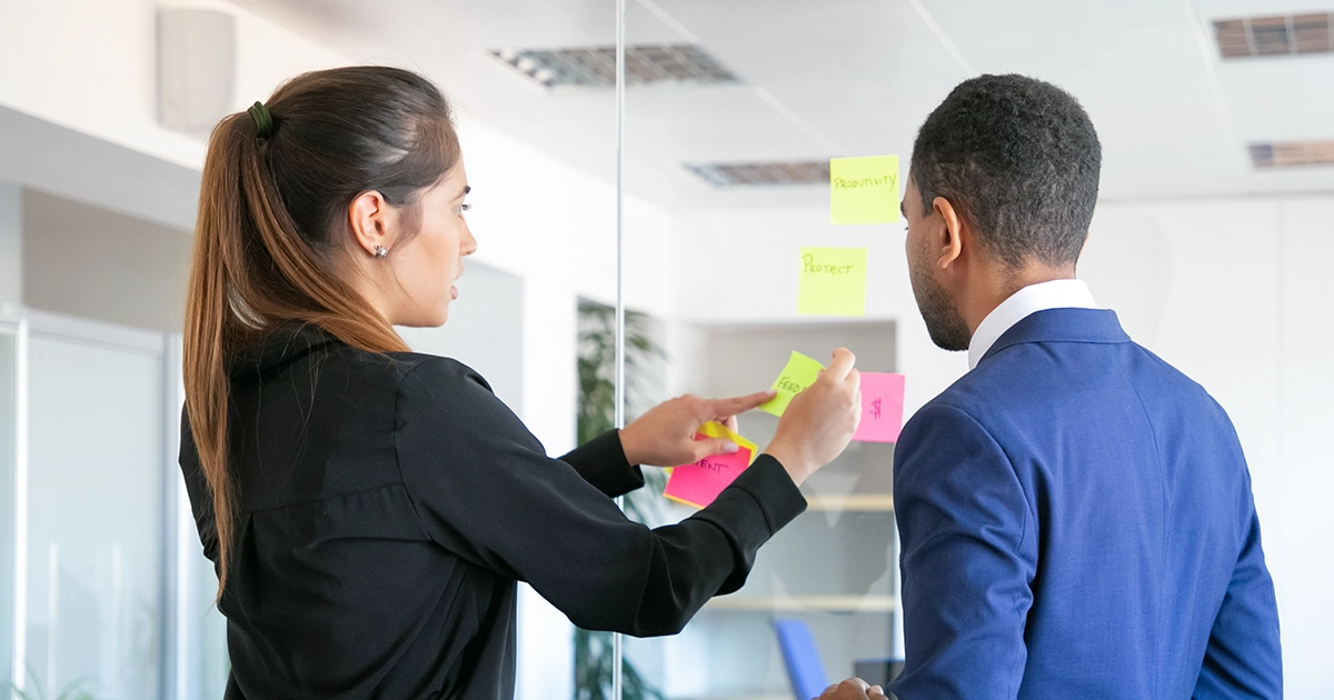 Duas pessoas de RH analisam post-its coloridos em uma parede de vidro, discutindo a classificação de talentos e como evitar o viés na avaliação de potencial no 9 box.