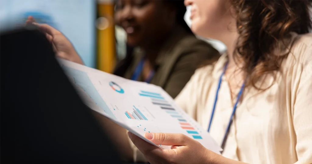 Líder e colaboradora analisam relatório impresso com gráficos e metas durante reunião de feedback e avaliação de desempenho na empresa.