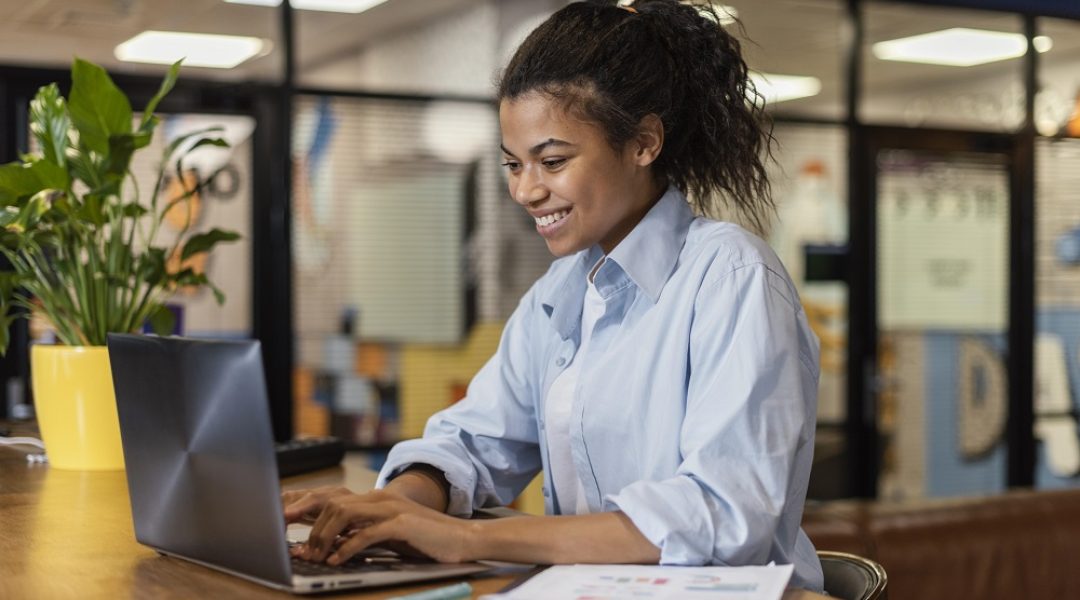 Na imagem há uma mulher preta com cabelo preso e uma blusa azul. Ela está sentada em frente a uma mesa com um computador e um vaso de plantas amarelho.