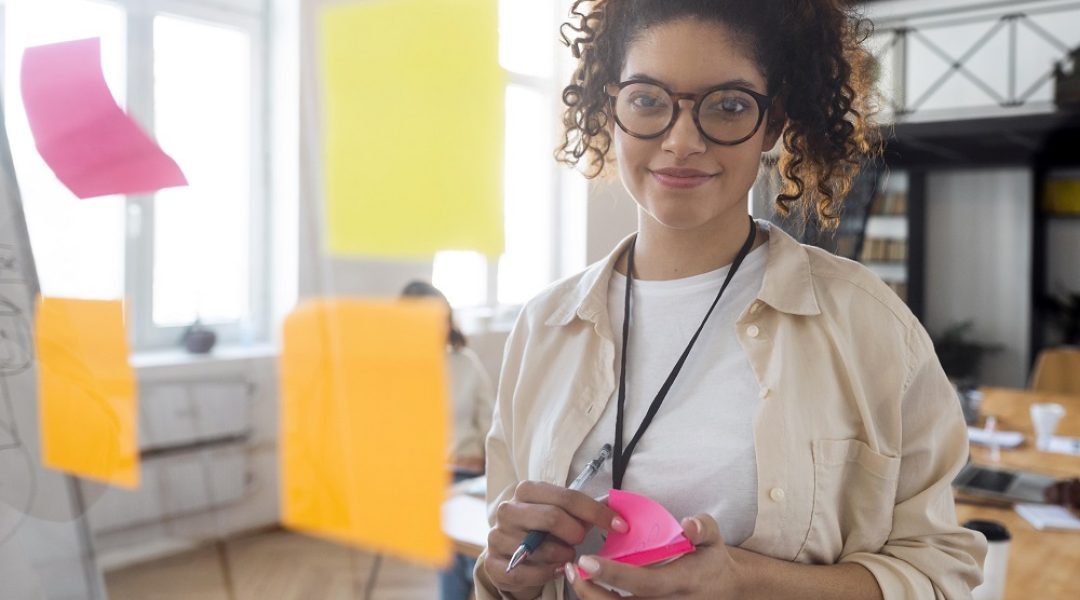 Na imagem há uma mulher sorrindo enquanto olha para frente de uma porta de vidro com quatro post-it colados