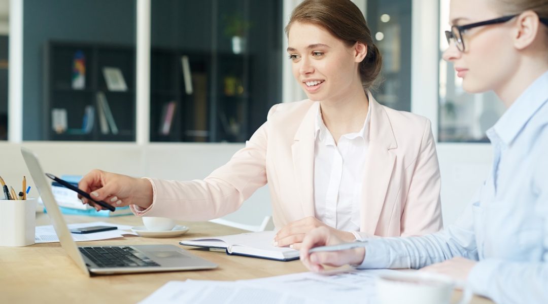 A imagem mostra duas mulheres de roupa social, elas estão olhando para um notebook em cima da mesa e uma delas está apontando a caneta para a tela.