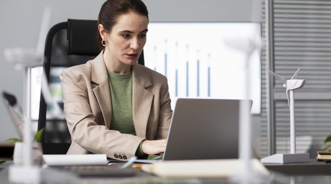 Uma mulher de cabelos presos está sentada em frete a um notebook. Ao fundo, existe um quadro com um gráfico de barras azuis, representando métricas essenciais para a gestão de desempenho.