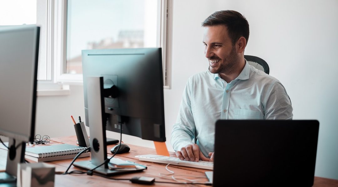 Homem sorridente trabalhando em um escritório, usando dois monitores. A imagem representa o uso da plataforma LMS no ambiente de trabalho.