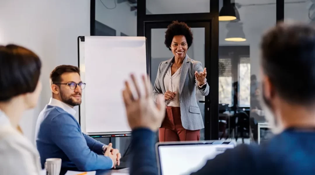 Quatro pessoas estão em uma sala de reunião, o foco está em uma mulher ao lado de um quadro em branco e um homem levantando a mão para fazer uma pergunta. A imagem representa uma conversa para o treinamento corporativo.