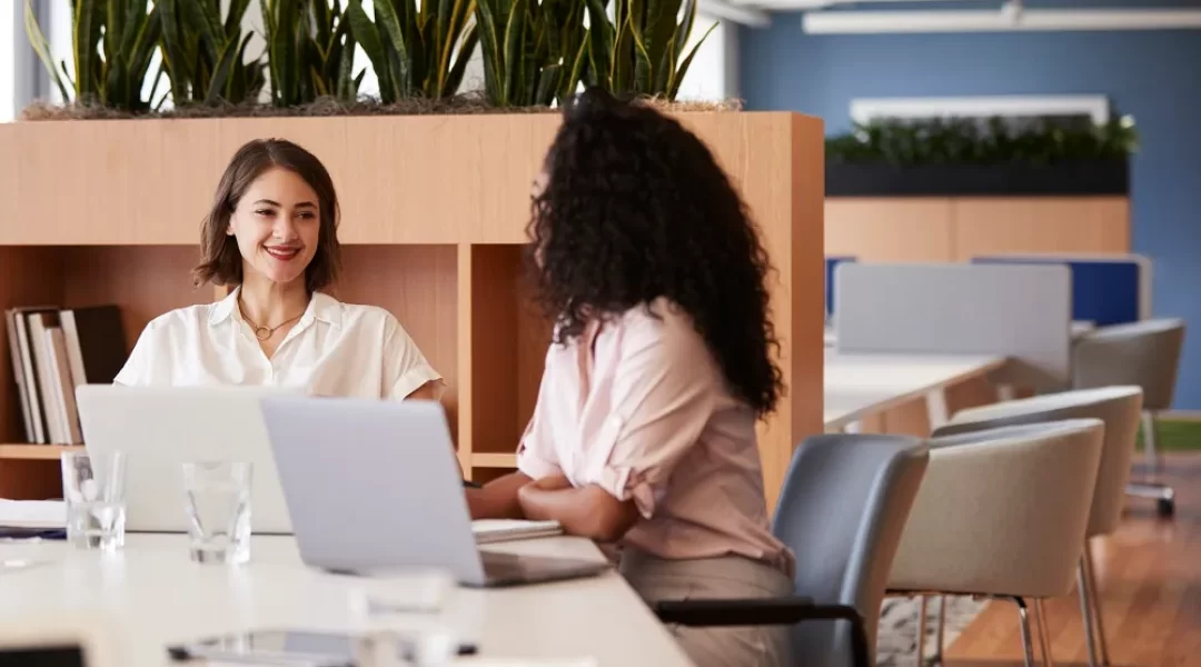 Duas mulheres estão sentadas em frente a um computador, uma delas está sorrindo enquanto olha para a outra. A imagem representa um dos tipos de feedback.