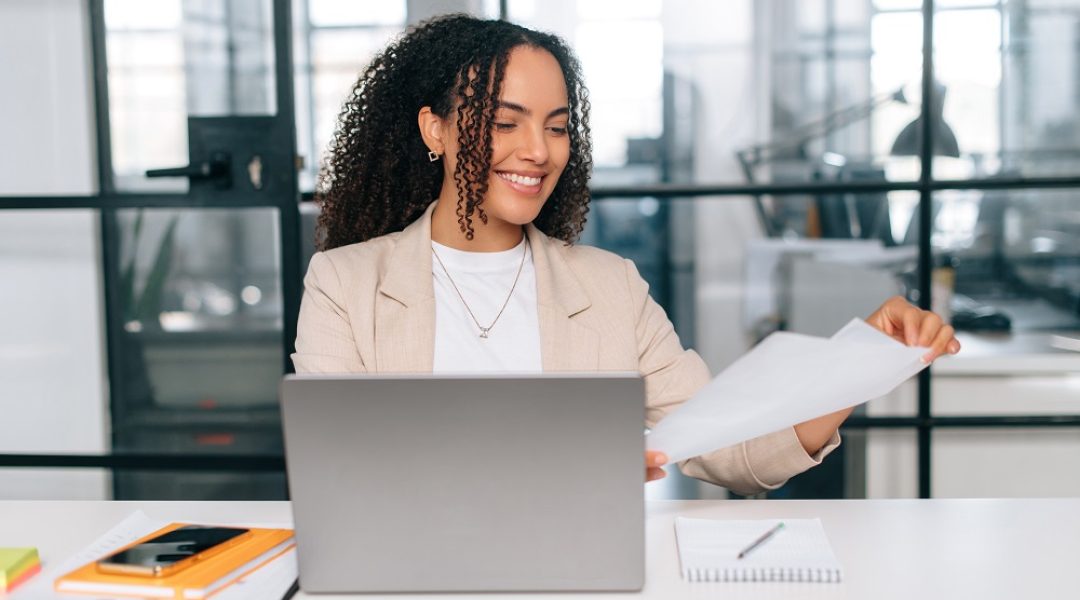 Mulher de cabelos cacheados está sorrindo enquanto olha alguns papéis em sua mão. A sua frente, há um notebook aberto e uma agenda laranja com um celular em cima.