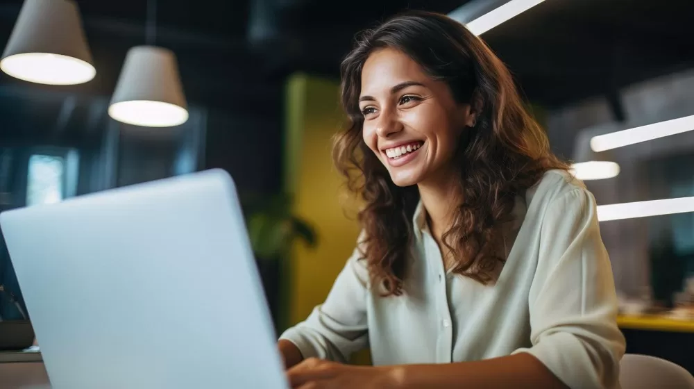 Mulher sorrindo para a tela de um notebook, ela está em um escritório. A imagem representa a satisfação de contar com a avaliação de desempenho.