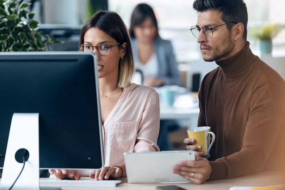 Um homem e uma mulher estão sentados em frente a um computador, ela está digitando no teclado enquanto ele segura um tablet em uma mão e uma caneca amarela e branca na outra. 