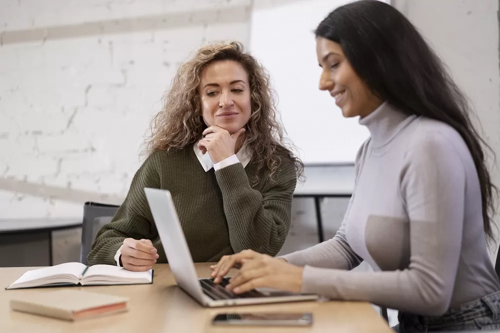 Duas mulheres estão em uma sala de reunião, uma está escrevendo no notebook enquanto a outra está com uma agenda. A imagem representa uma reunião de avaliação de desempenho.