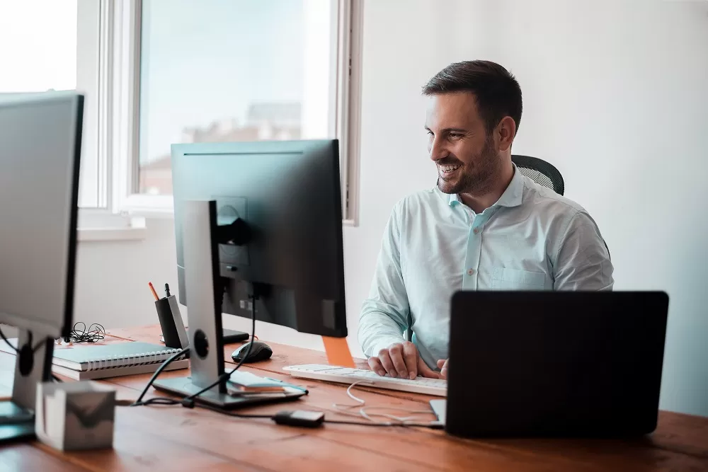 Homem sorridente trabalhando em um escritório, usando dois monitores. A imagem representa o uso da plataforma LMS no ambiente de trabalho.