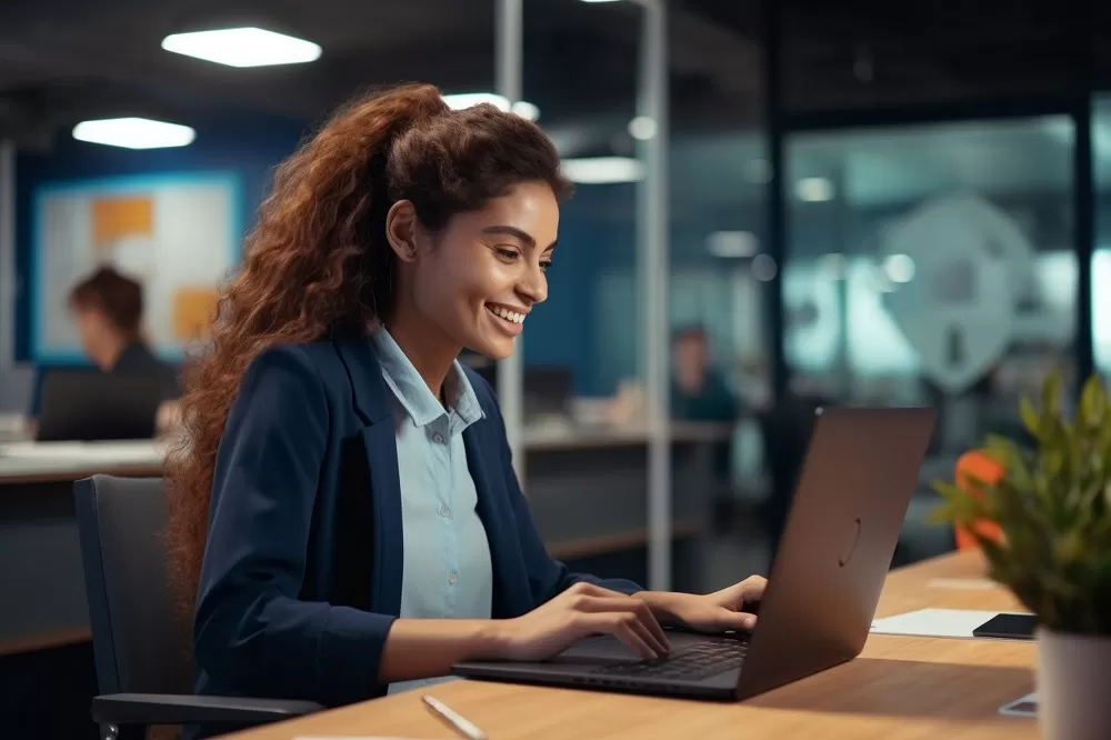 Mulher de cabelos castanhos e cacheados está sentada em frente a um computador. Ao fundo, existem outras pessoas embaçadas para dar foco em quem está na frente.