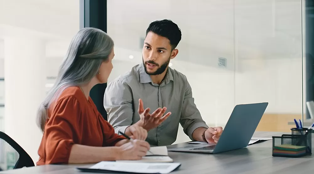 Um homem e uma mulher estão em uma reunião, o foco está no homem que conversa em frente a um notebook aberto.
