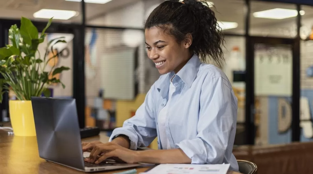 Na imagem há uma mulher preta com cabelo preso e uma blusa azul. Ela está sentada em frente a uma mesa com um computador e um vaso de plantas amarelho.